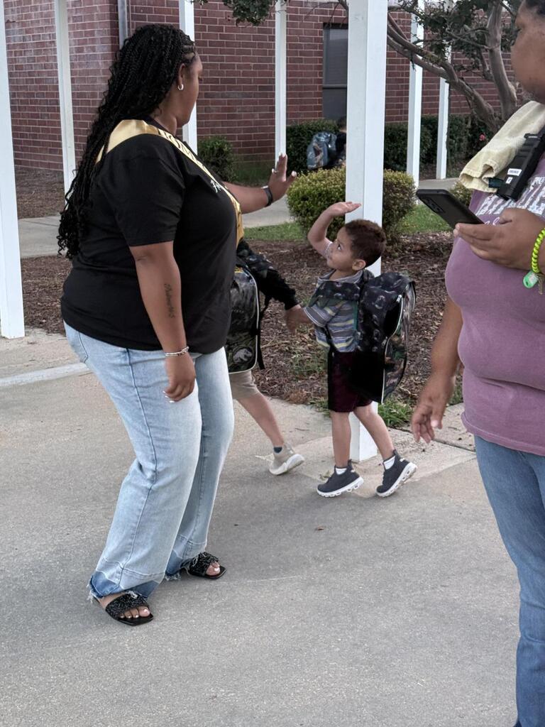 A woman stands outside near a building with brick columns. Children, one in a backpack, are walking away.
