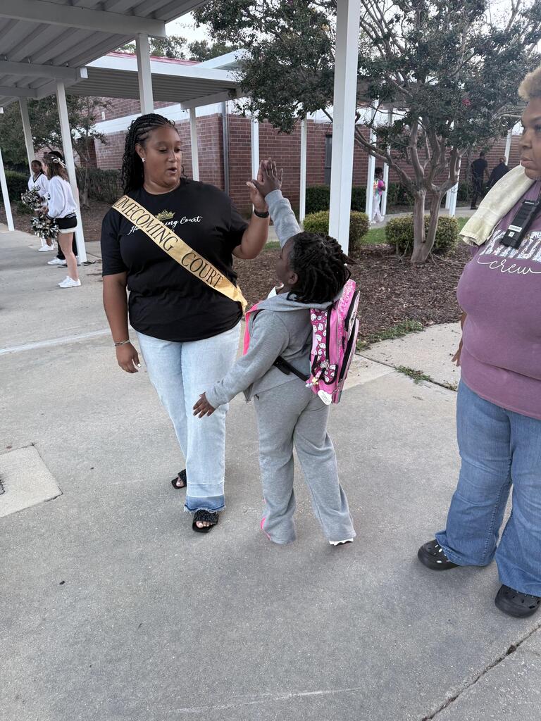 A woman wearing a sash and sneakers helps a girl with a pink backpack on a concrete walkway.