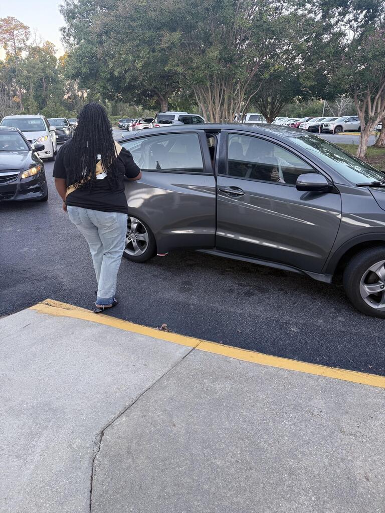 Person standing by a parked gray Honda in a parking lot with other vehicles and trees.