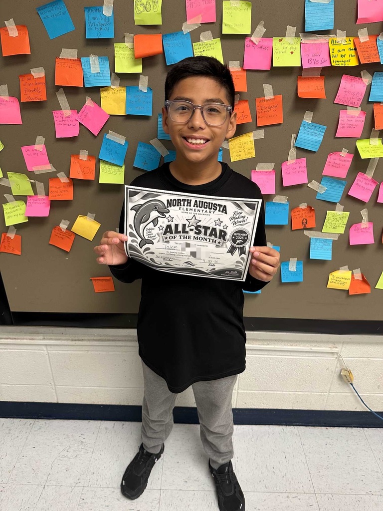 Child in a black shirt holds a certificate with a fish illustration, smiling. Colorful sticky notes cover the background wall.