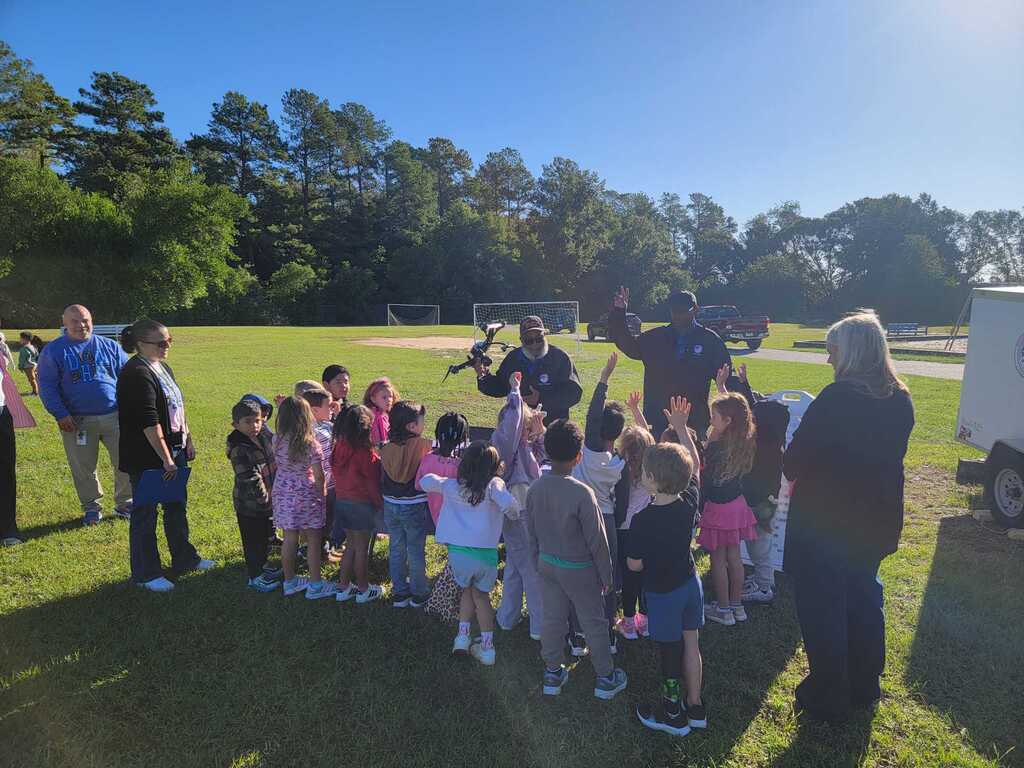 Children and adults stand in a circle on a field, with one person holding a microphone. Green trees are in the background.