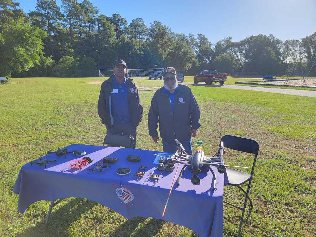 Two individuals stand next to a table with a blue cover, displaying various equipment, in an open grassy field.