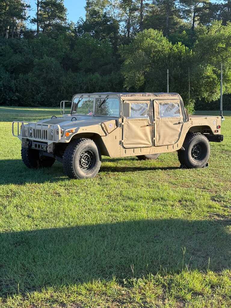 A beige military vehicle parked on grass. Trees and shrubs are visible in the background.