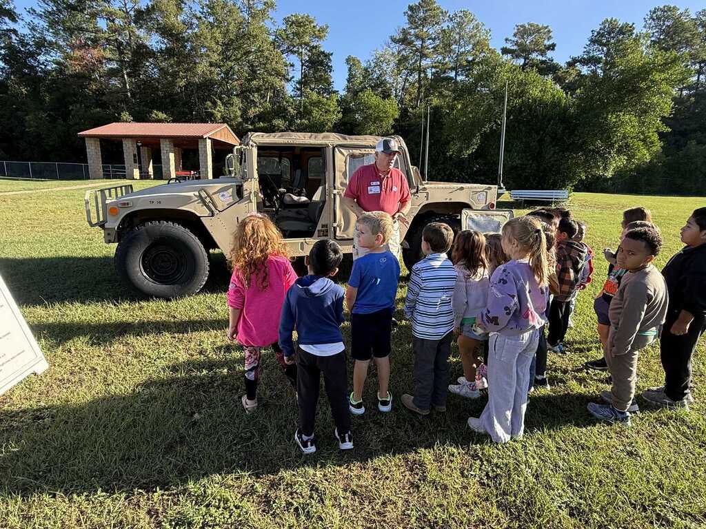 Kids gathered around an armored military vehicle with a man standing in front of it on a grassy field.