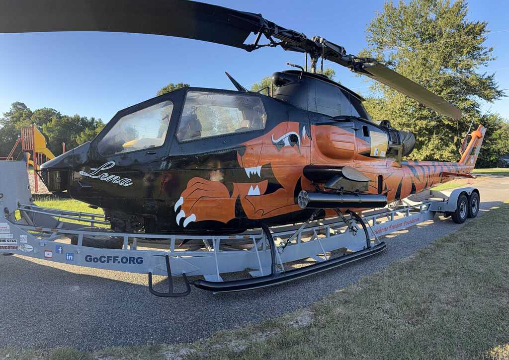 Helicopter with black and orange design on a trailer on a grassy surface. Trees in the background.