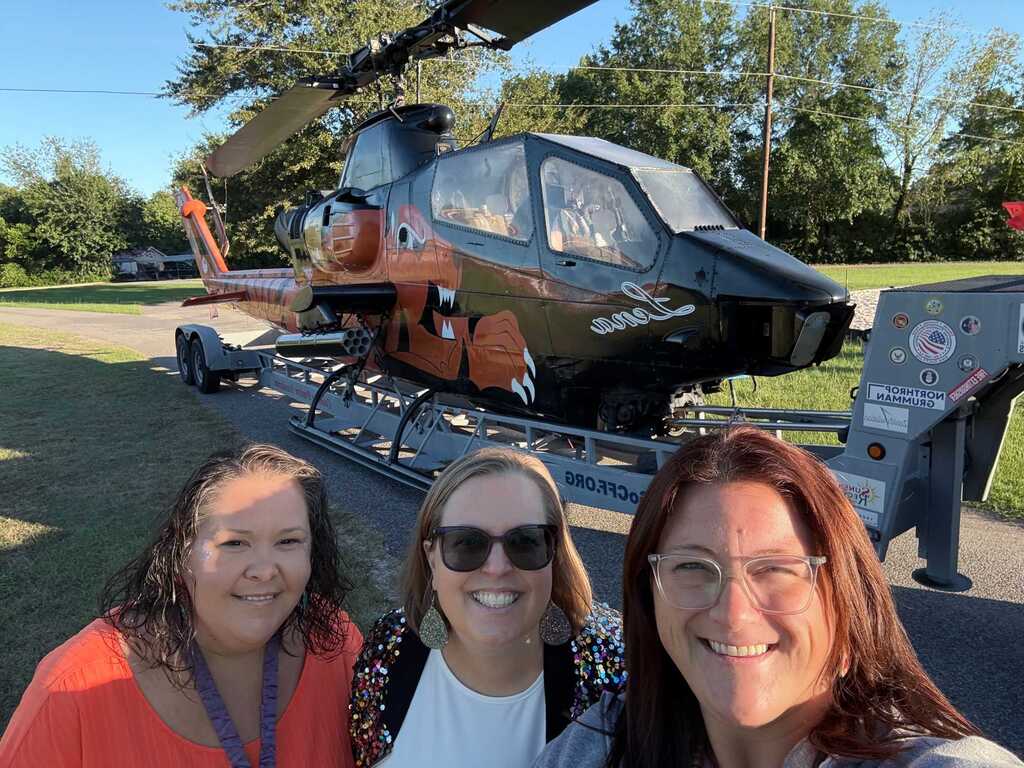 Three women smile in front of a black and orange helicopter parked on a platform.