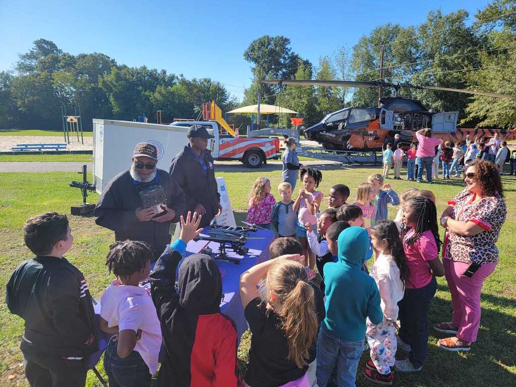 Group of children and adults outdoors, with a helicopter in the background. A table holds objects in front of them.