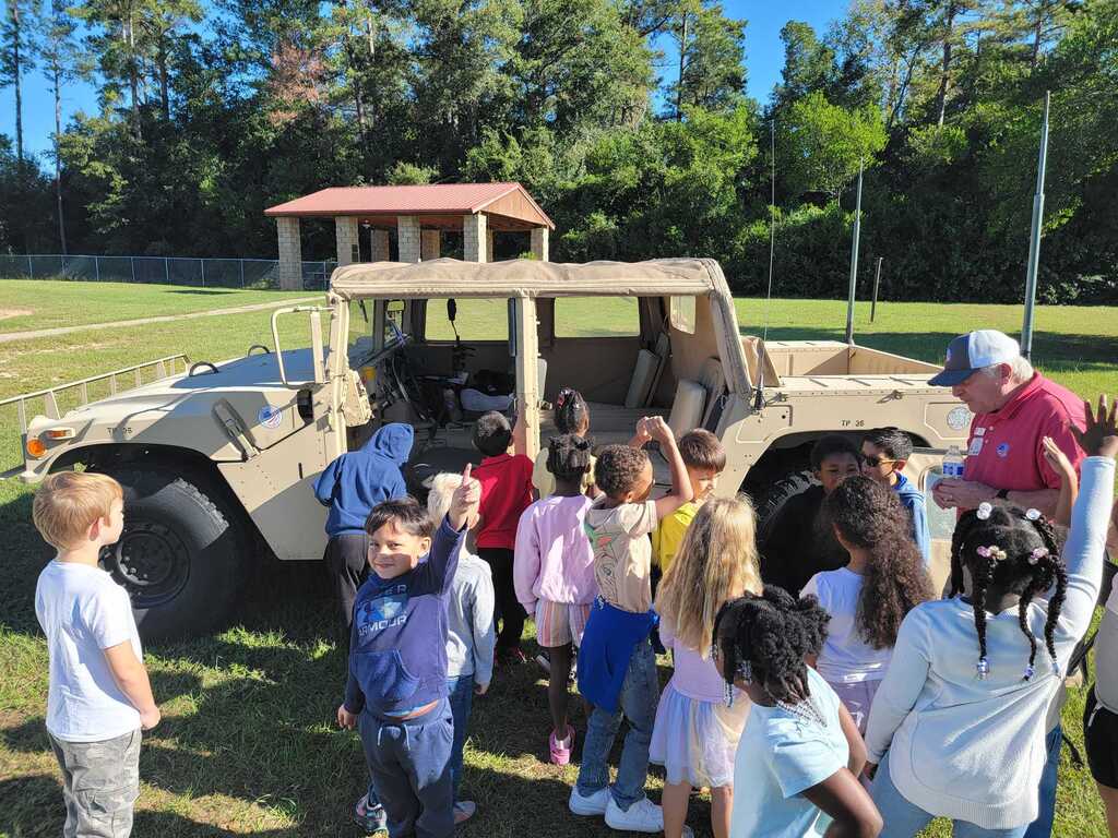 Kids stand around a military vehicle with a red roof. A man stands in front, raising his hand. Trees and grass surround them.