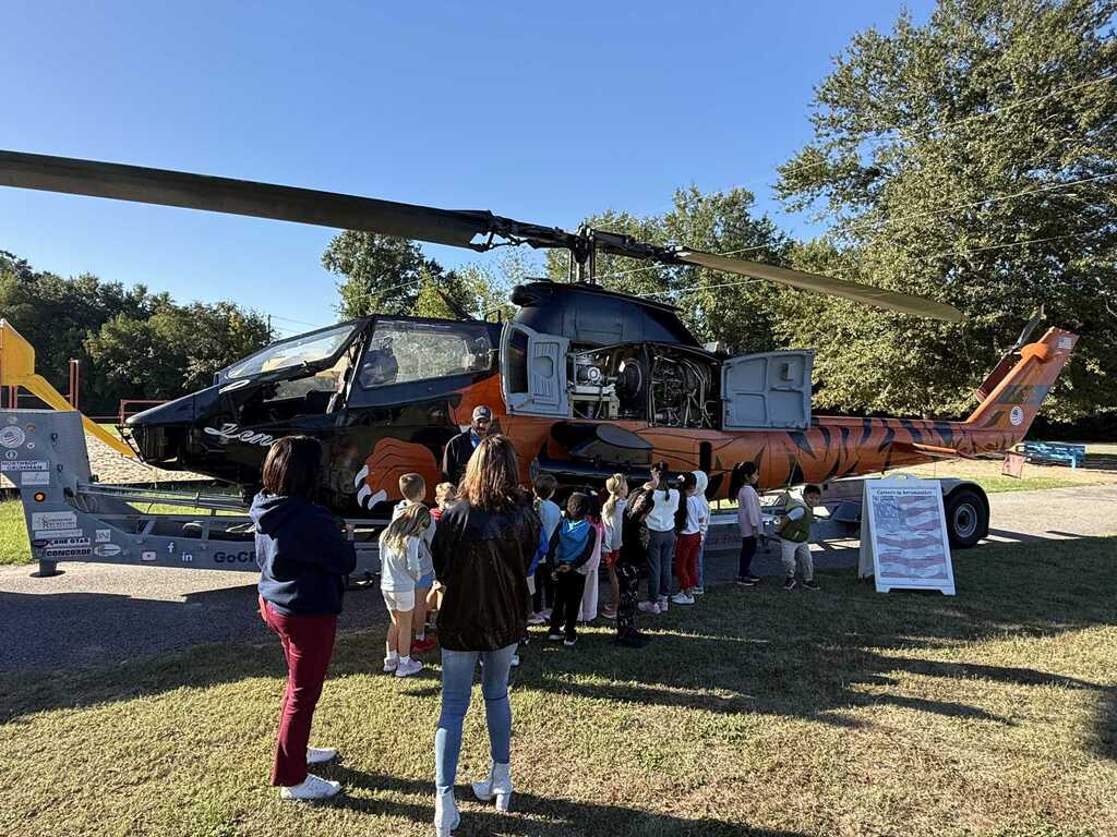 A group of people stands around an orange and black helicopter. Trees are in the background.