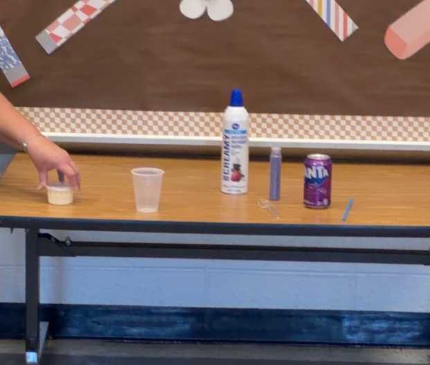 A hand reaches for a small cup on a table. Nearby are a small cup, a bottle, a can, and a tube.