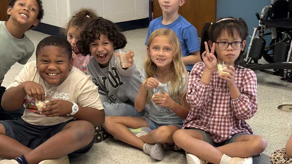 A group of children seated on the floor, smiling, with one holding a peace sign.