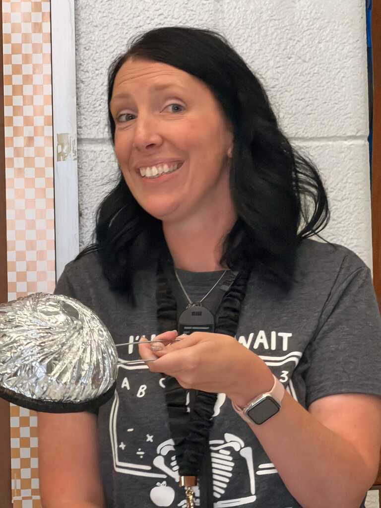 A woman in a gray T-shirt holds a silver object while smiling, standing next to a white-and-brown wall.