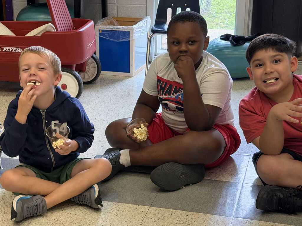 Three kids eating snacks; one holds a banana, another has a sandwich, and the third holds a cookie.