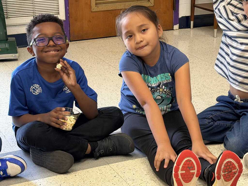 Two children sitting on a floor, one eating and the other smiling. Both wear blue shirts, black pants, and sneakers. Background shows a tiled floor.