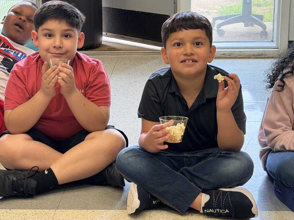 Two boys seated, one with food in hand, another with a drink, against a blurred background.