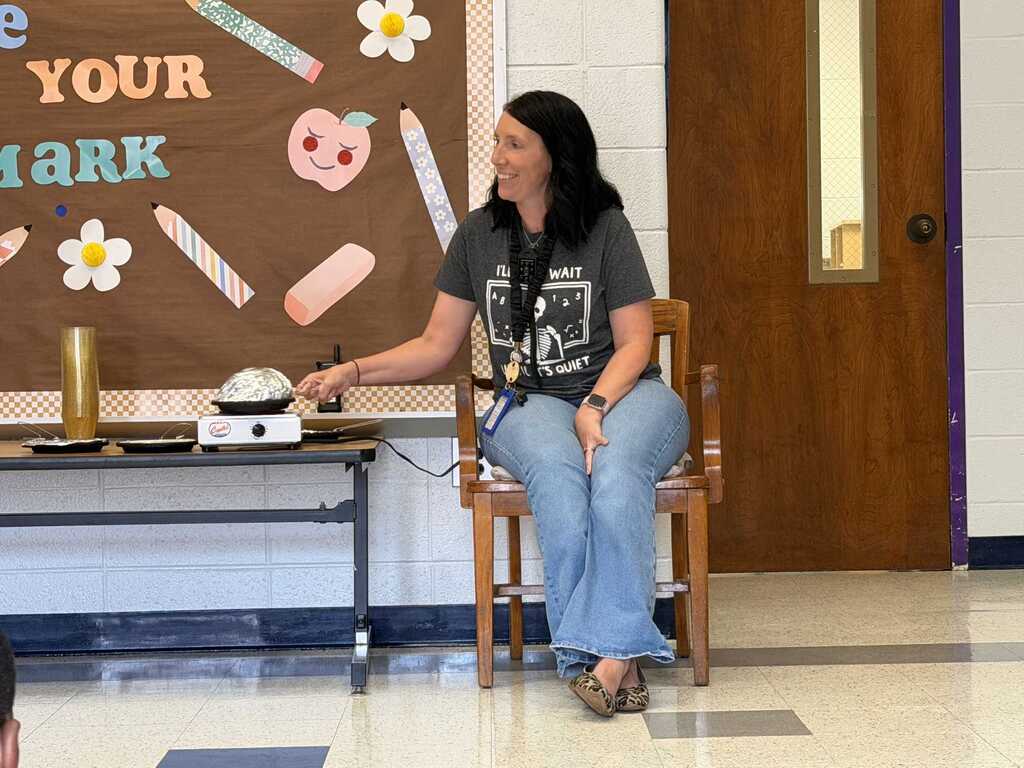 A woman sits in a chair before a table, in front of a classroom door. Behind her, a bulletin board displays "Make Your Mark" with colorful designs.