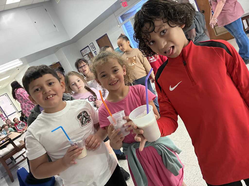Three kids standing together, smiling and holding cups with straws. One wears a red jacket, another a pink dress, and the third a white shirt.