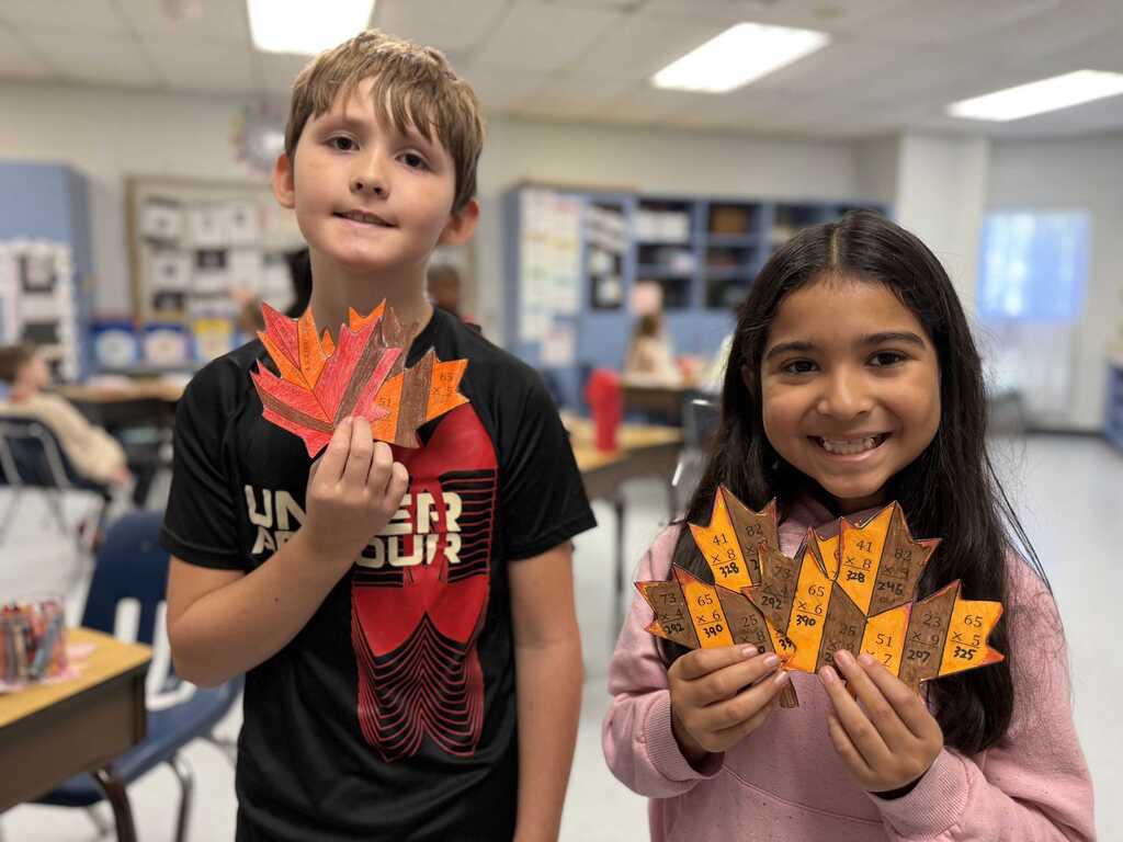 Two children, a boy and a girl, hold up decorated leaves, smiling at the camera. Background: classroom with tables, chairs, and a whiteboard.