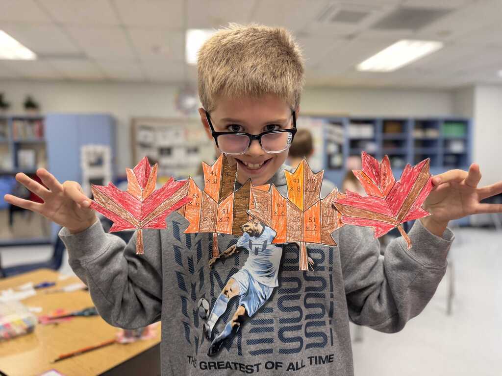A boy wearing glasses and a gray sweatshirt holds a colorful paper art piece in both hands in a classroom.