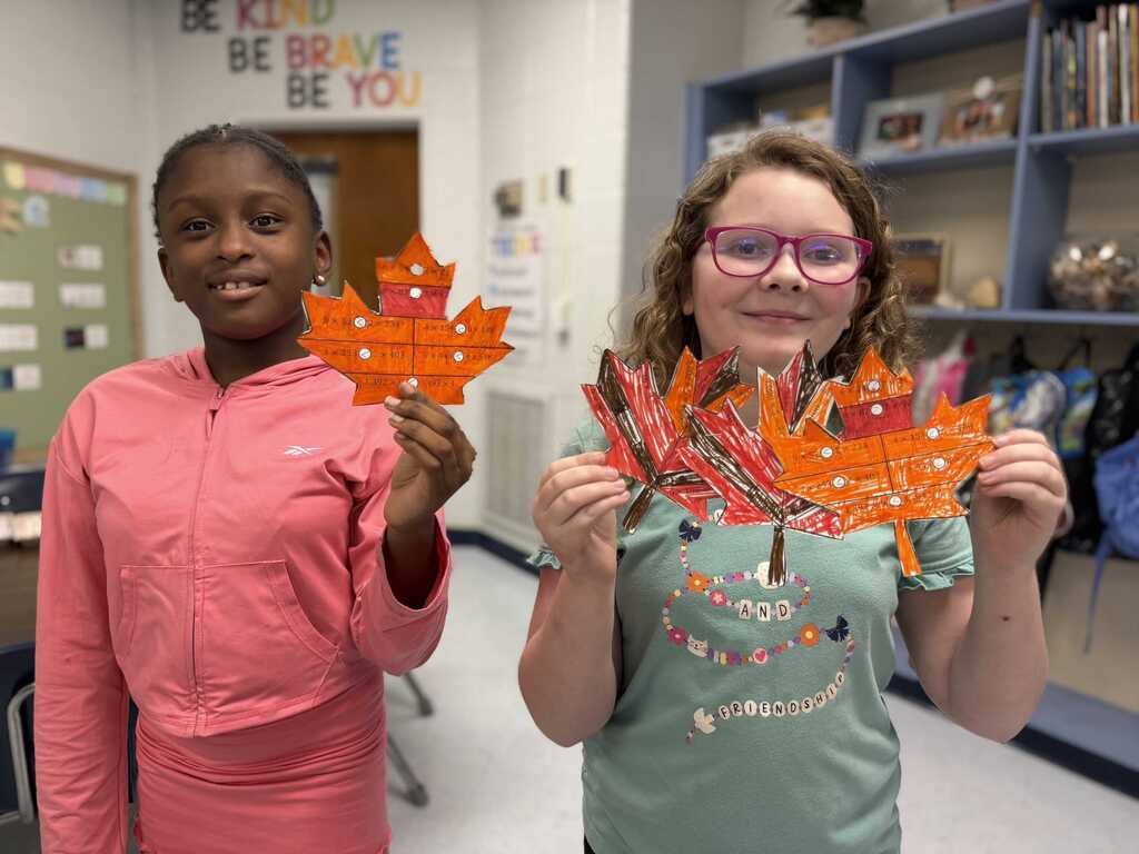 Two girls stand in a classroom, holding orange and yellow maple leaf art. One wears pink, the other green. Behind them, a bulletin board and shelves with books are visible.
