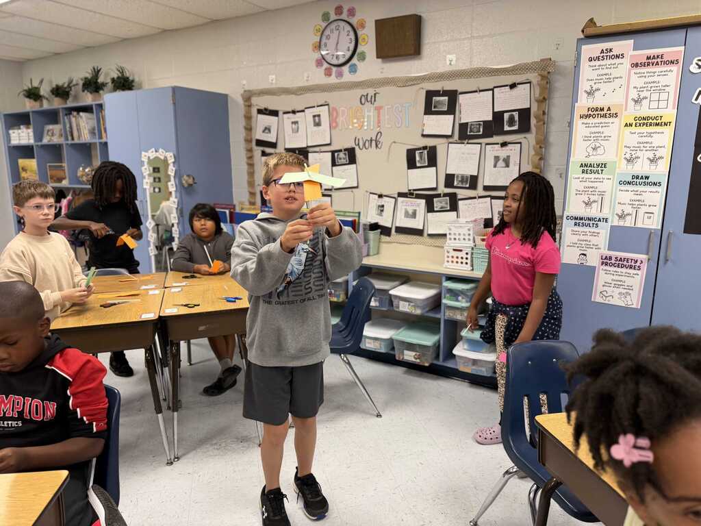 A classroom with children seated at tables. A boy stands holding a paper object. A woman stands behind him.