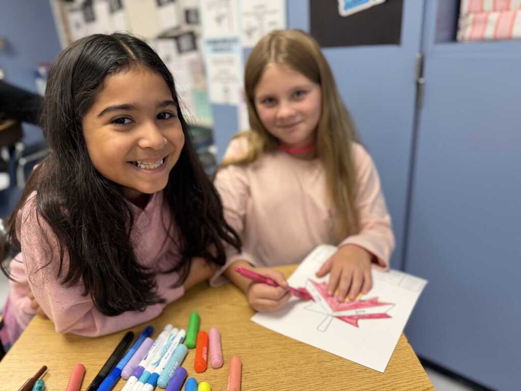 Two girls in a classroom, seated at a table. One smiles, the other cuts a paper. Various colored markers on the table. Blue walls in the background.
