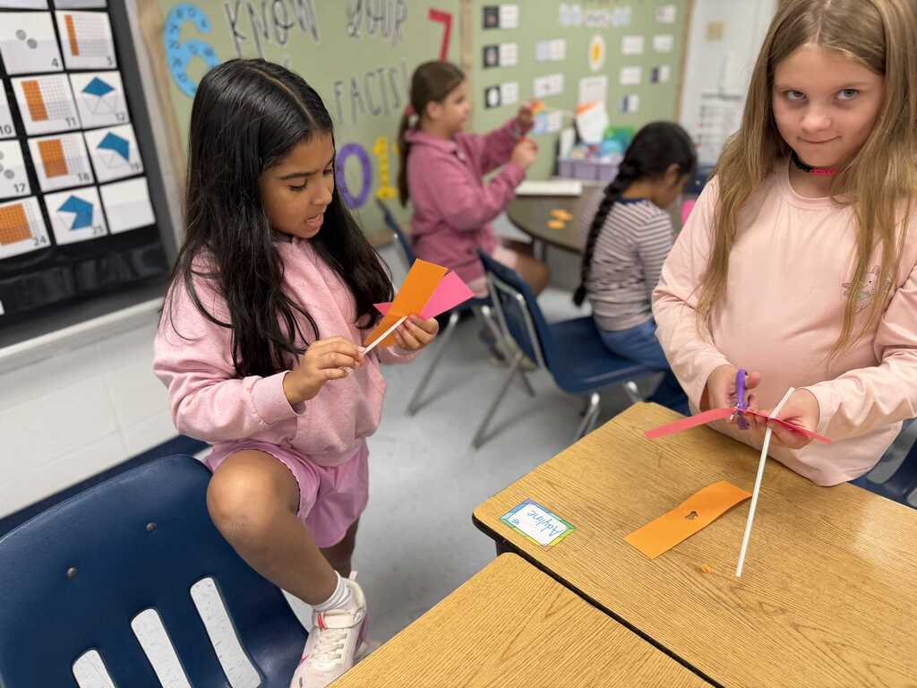 Two girls in pink shirts work on a project at a desk in a classroom with other students.
