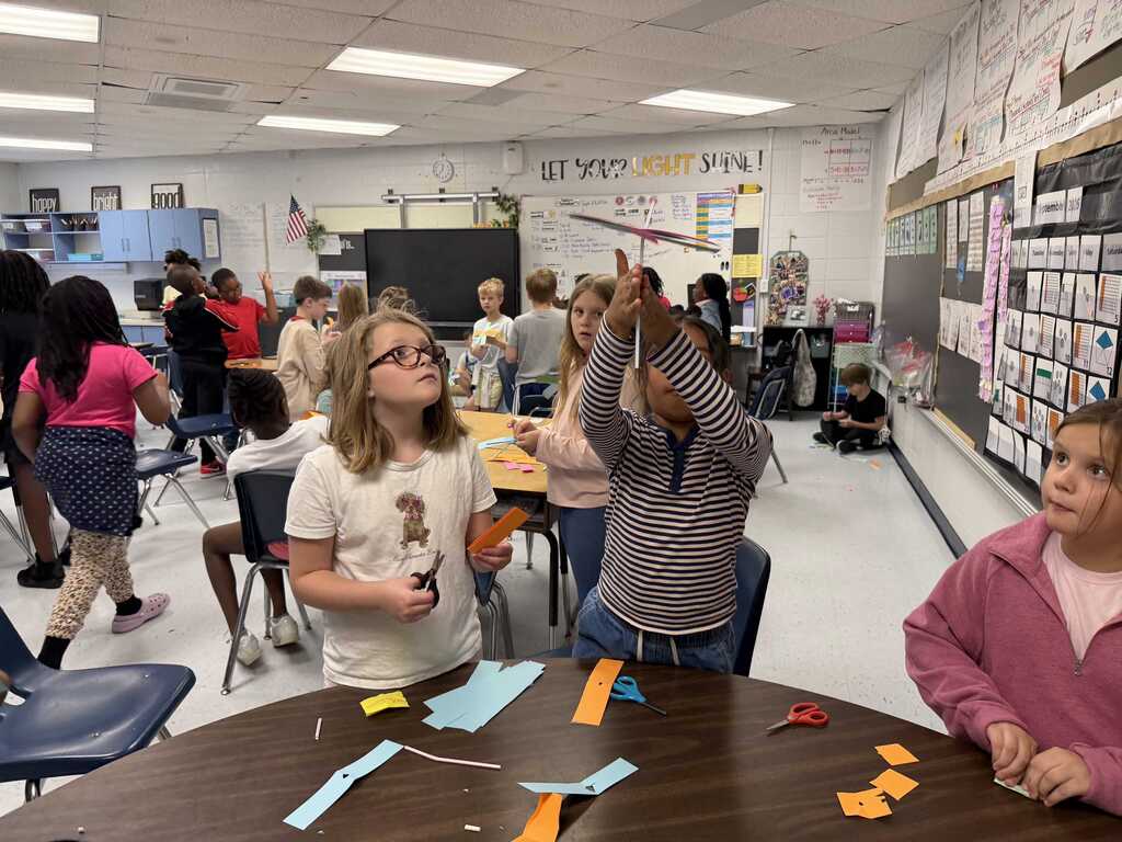 A classroom with students standing around a table with paper. Some are raising their hands.