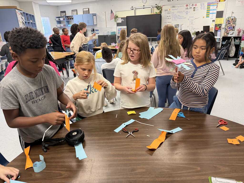 A classroom with students gathered around a table. They hold colored papers, scissors, and pencils. Some are seated and others stand.