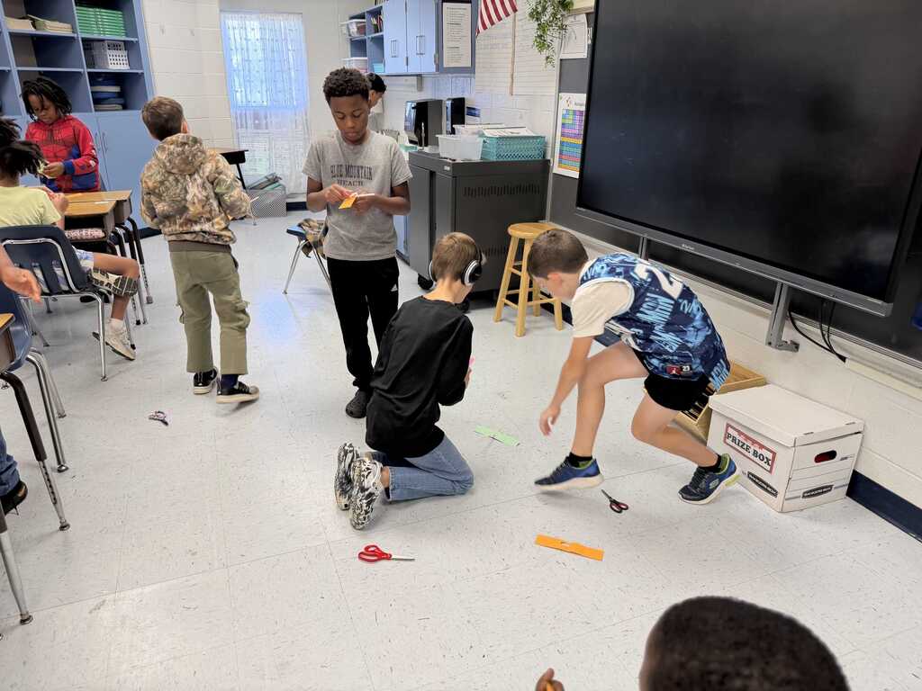 Several students in a classroom, some kneeling and interacting with items on the floor, with a large monitor on a stand behind them.