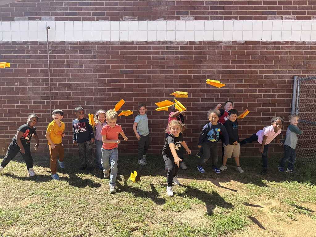 A group of kids holding yellow objects in front of a brick wall on a grassy area.