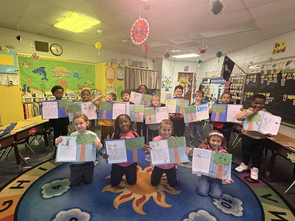 Group of children holding up papers in a classroom with colorful rugs and desks.
