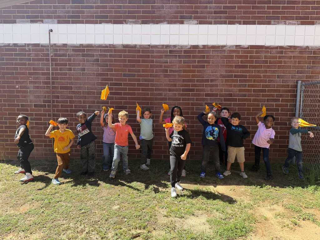 A group of children wearing gloves holding yellow objects stand in a line against a brick wall.