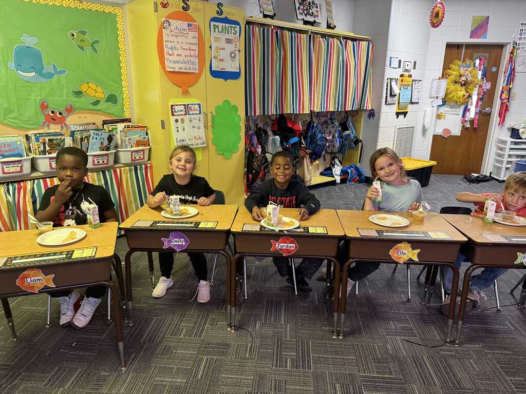 Children sit at desks with plates, smiling, in a classroom with colorful walls, posters, and a door.