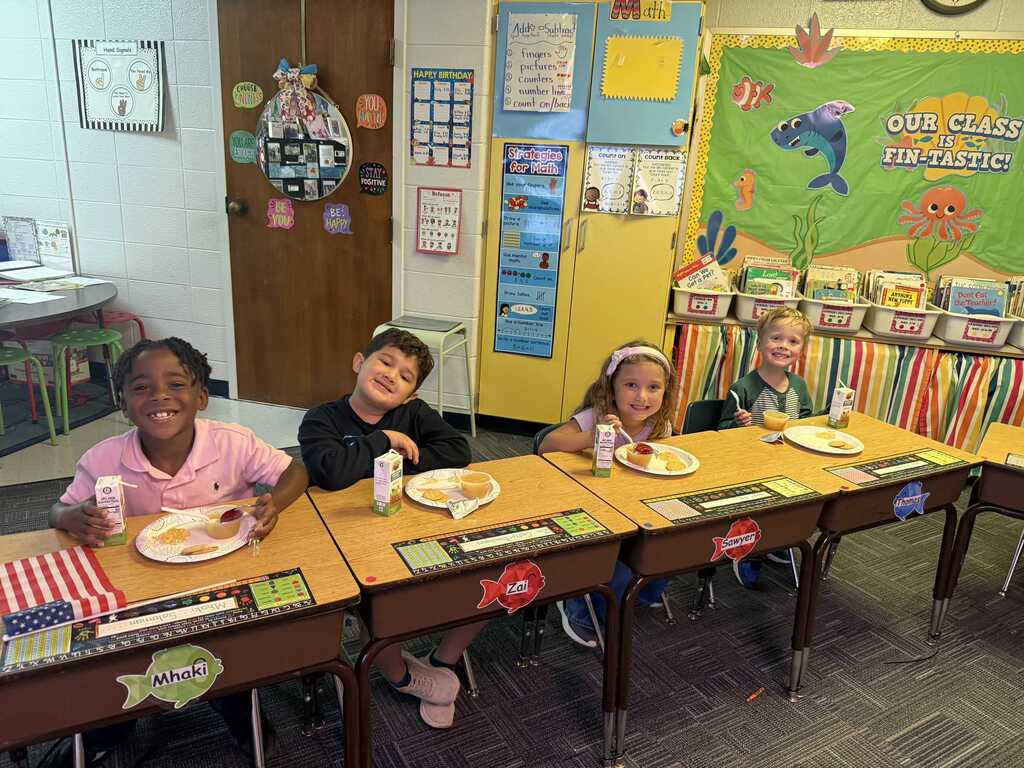 Four children seated at a classroom table, eating with plates in front of them. Brightly colored classroom decor on the walls.