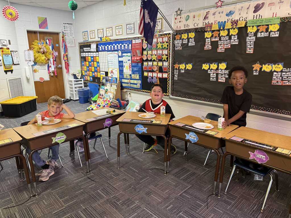 Three children seated at desks in a classroom with a bulletin board and blackboard in the background.
