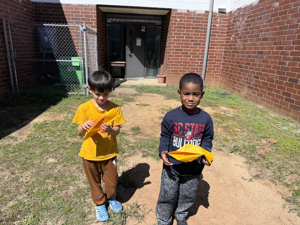 Two children in front of a building with brick walls. One holds a yellow item.
