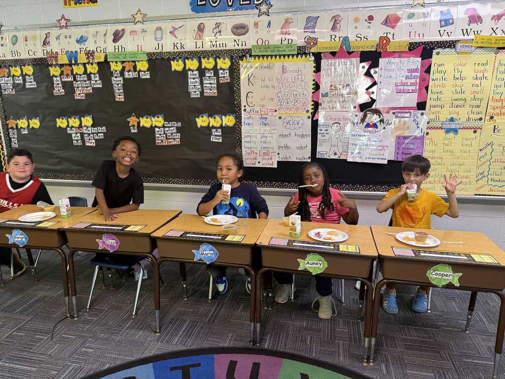 Five children seated at desks, facing the camera, holding cups and eating. Behind them is a bulletin board with papers.