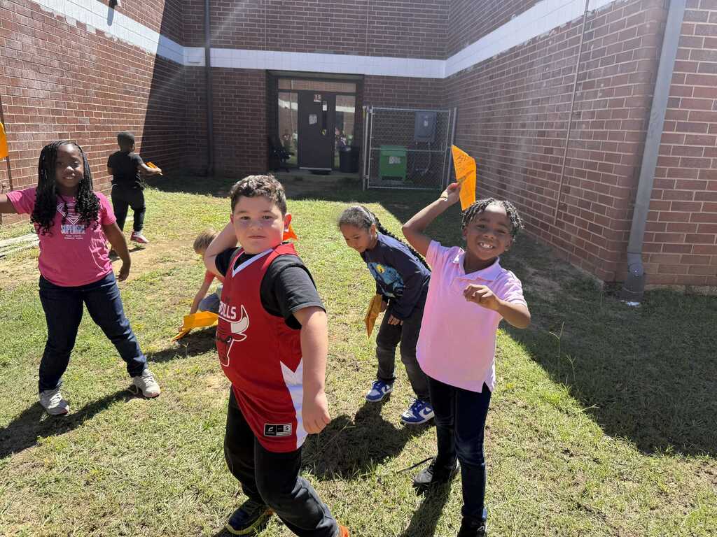 Children playing outdoors. A brick building in the background. A girl in pink and boy in red raising yellow objects.