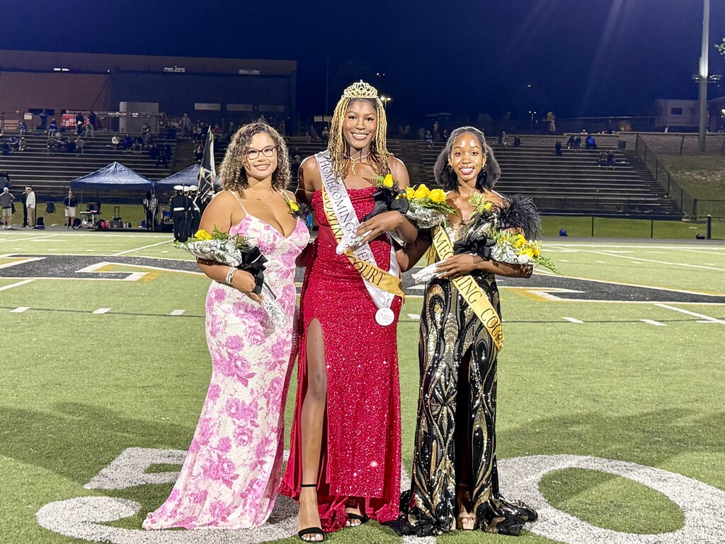 Three women stand on a football field at night, each holding flowers. The woman in the middle wears a sash.