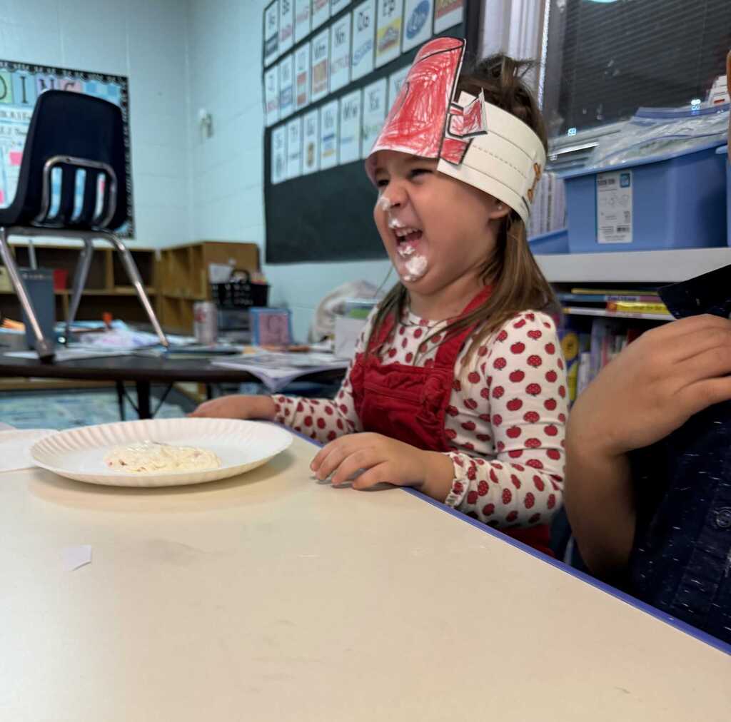 Child in a red apron sits at a table, wearing a red hat. A paper plate with food sits before the child.