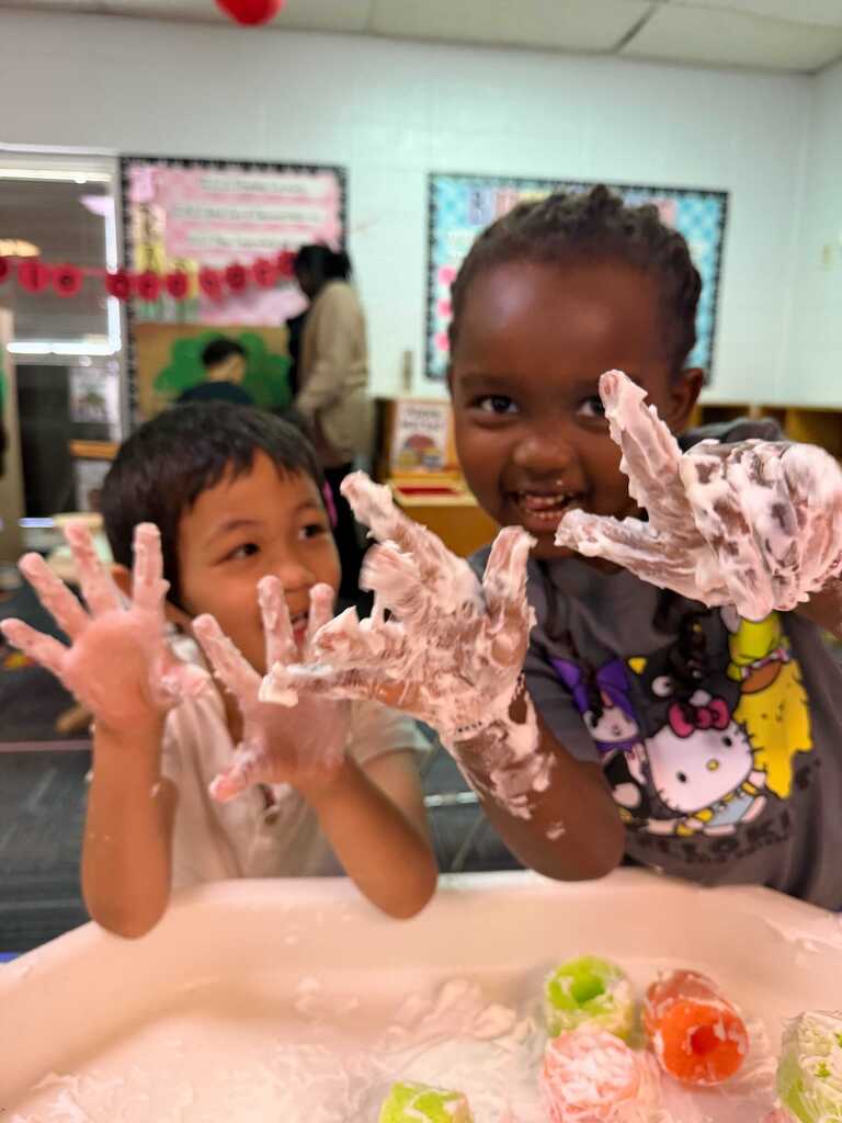 Two kids sit before a table, one with hands covered in soap, the other smiling. The table holds colorful objects.