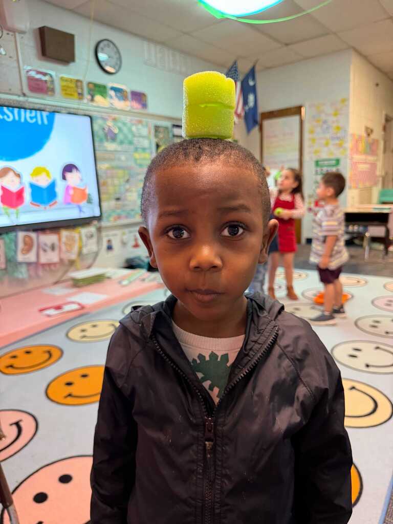 A child with a yellow cylinder on his head stands in a classroom with smiling faces on the floor.