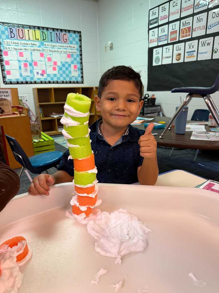 A young boy holds a colorful tower of foam pieces while sitting in a classroom.