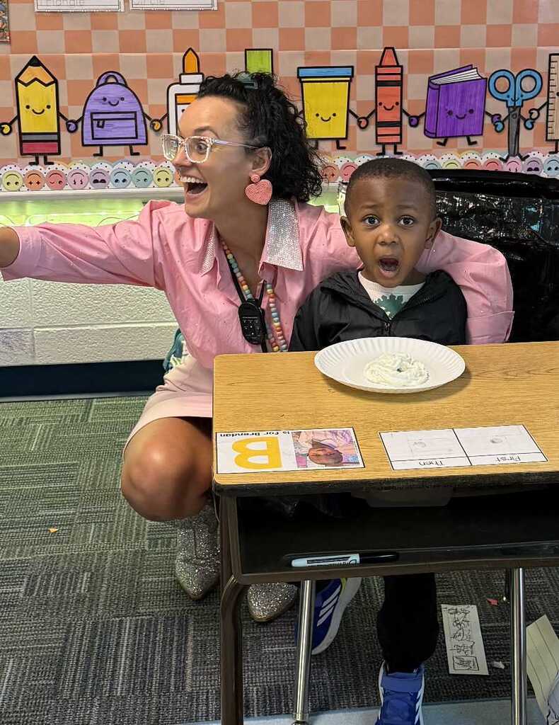 A woman in pink kneels, holding a boy in a black jacket. They share a laugh. Behind them, a wall displays colorful graphics and a desk holds a plate.