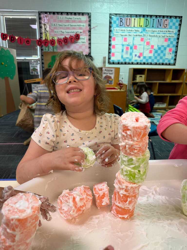 A young girl in glasses plays with colorful, cylindrical objects on a tray in a classroom.