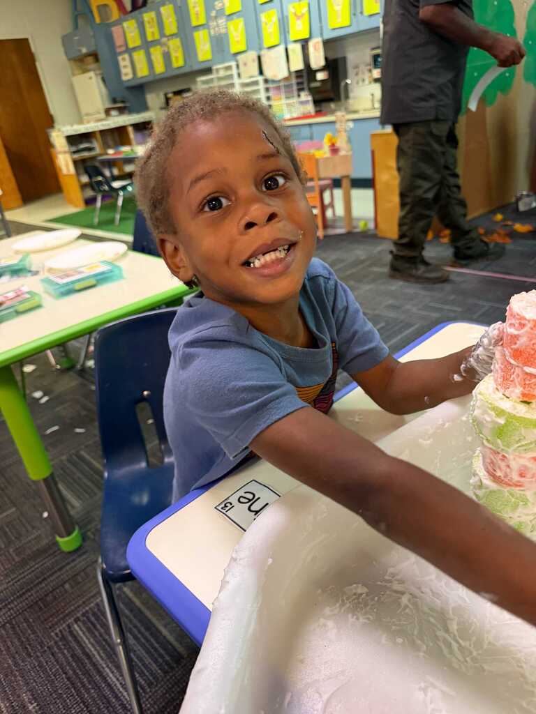 A young boy sits at a table, reaching for a cake. Behind him, a person stands near a table with various items.
