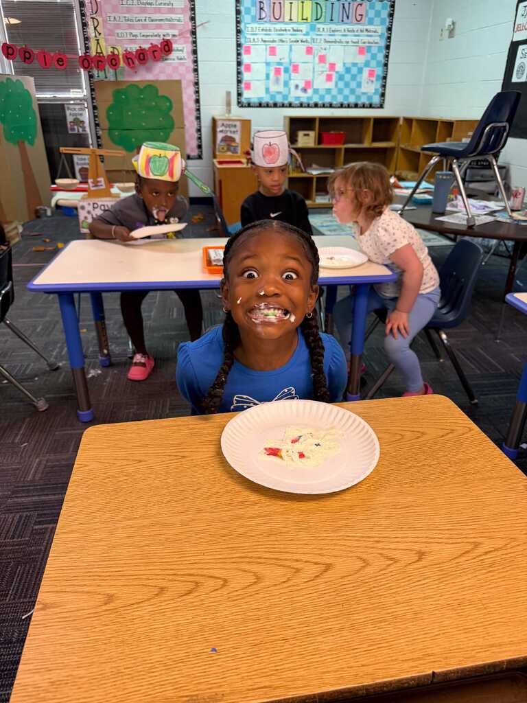 A classroom with tables and chairs. A girl with a surprised expression holds a paper plate.