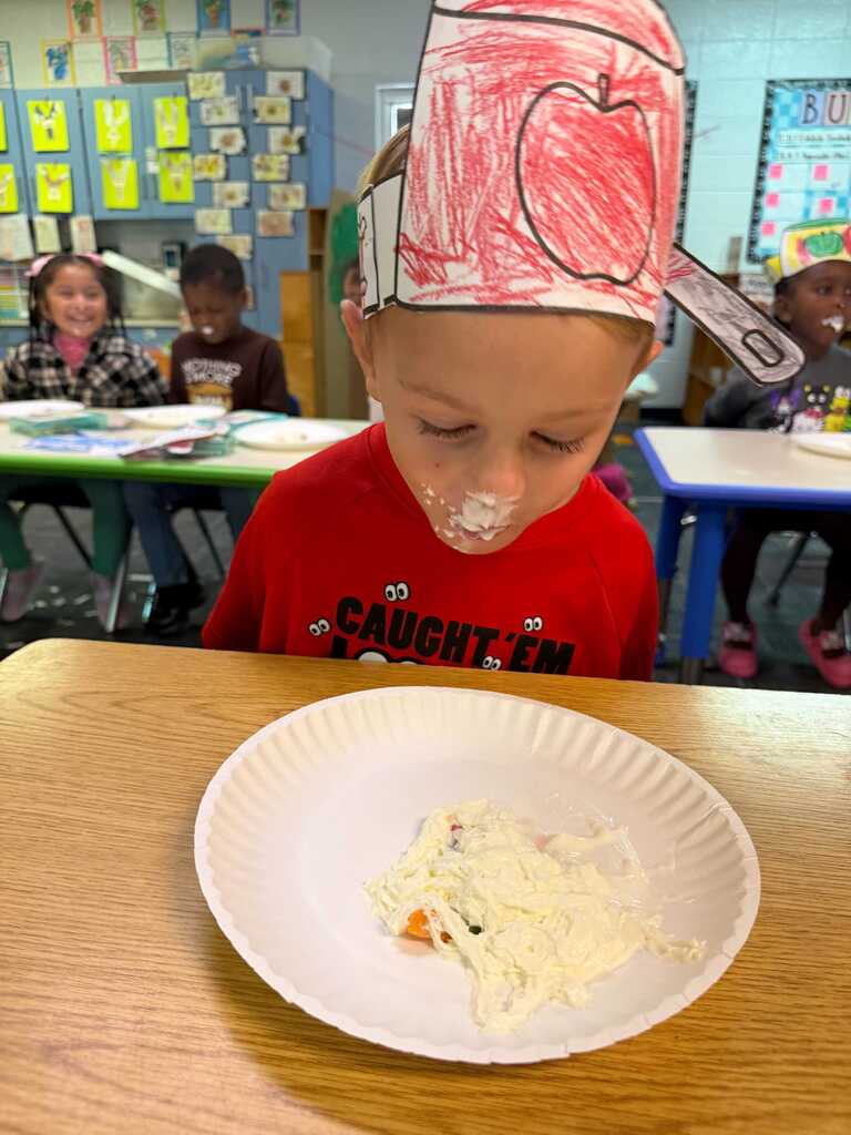 A child in a red shirt wearing a paper hat shaped like an apple eats a plate of food.