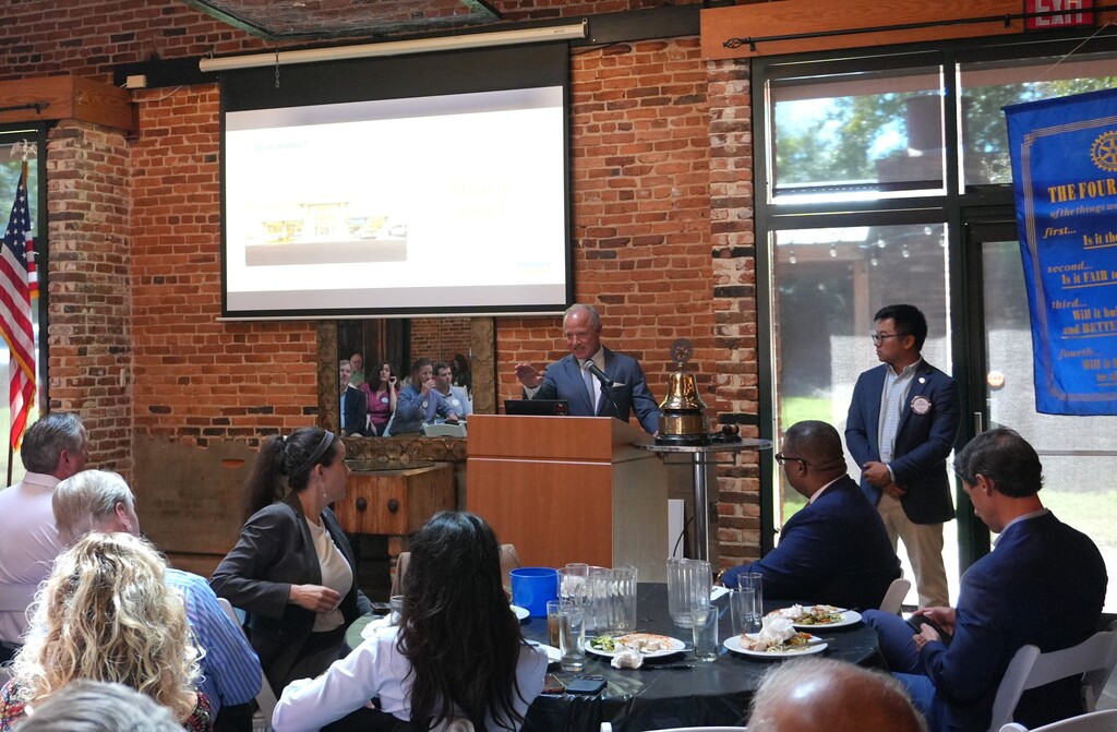 A man speaks at a podium with a projector screen in the background. People sit around tables.